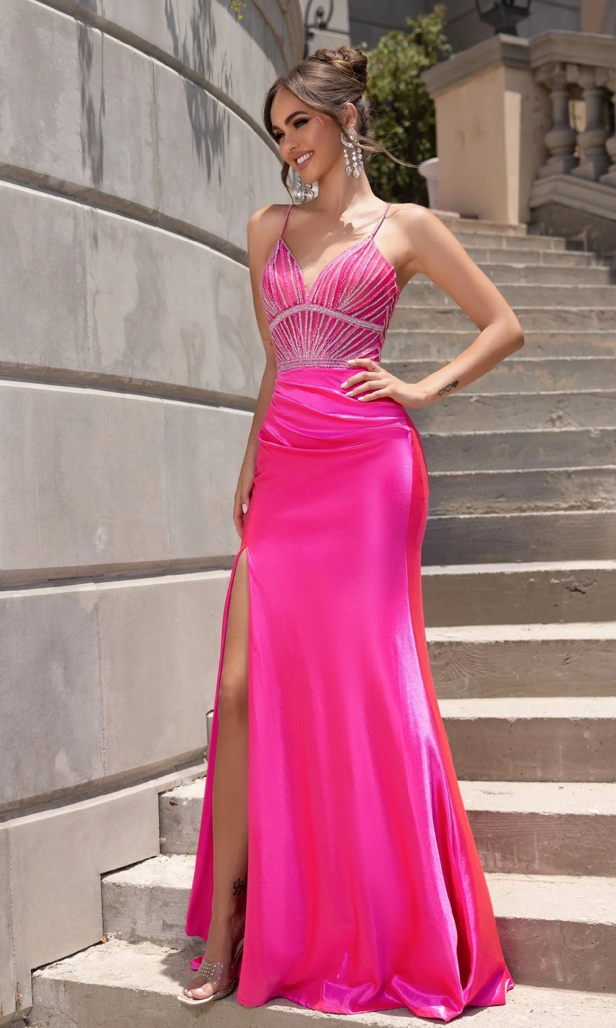 A woman smiles on outdoor steps in the Portia and Scarlett PS25244, a vibrant pink long prom dress featuring a beaded silver-embellished bodice, thin straps, sleeveless design, and a thigh-high slit.